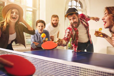Party playing ping pong with drinks and man in hat hitting ping pong ball with forehand